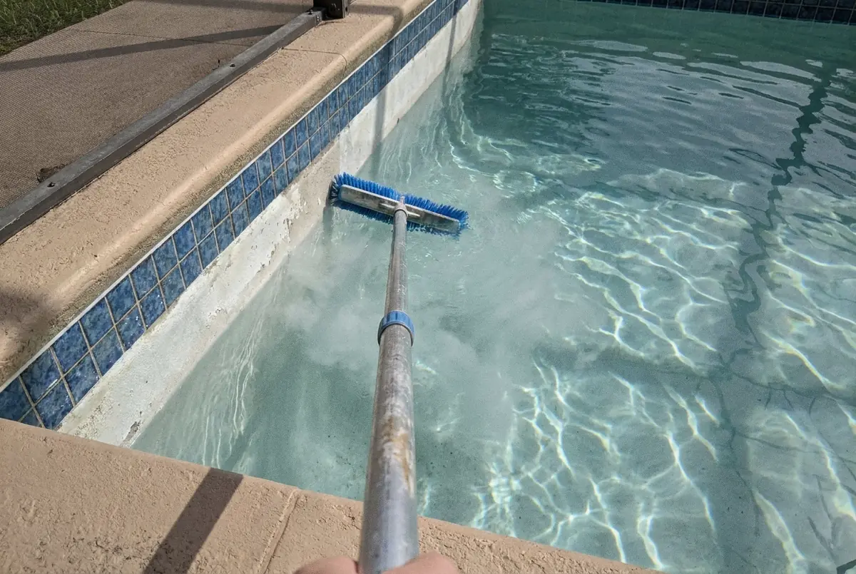 A pool technician brushing the tiled waterline of a clean blue pool with a telescopic pole